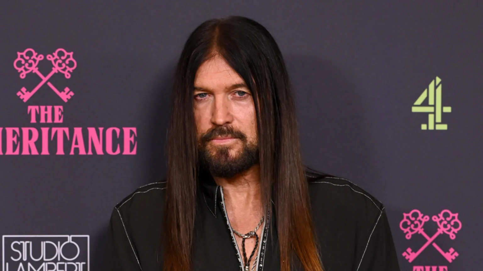 Billy Ray Cyrus, with long dark hair and a beard, looks forward, standing in front of a dark grey backdrop with "THE INHERITANCE" and "4" logos.