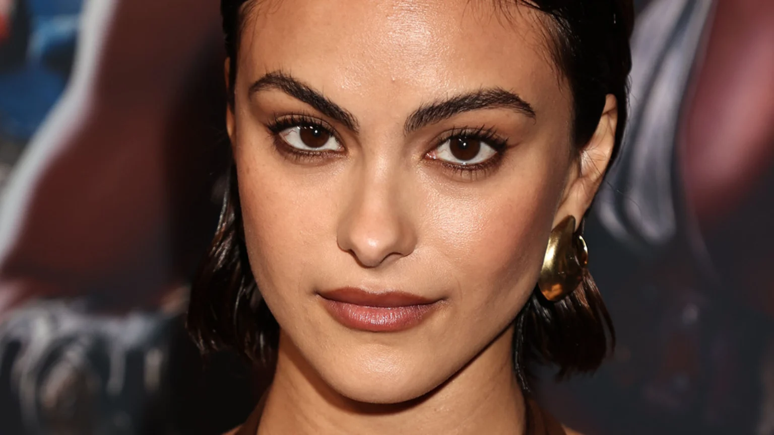 Close-up portrait of a woman with dark brown eyes, dark hair, defined eyebrows, subtle makeup, and a large gold-toned earring, looking directly forward.