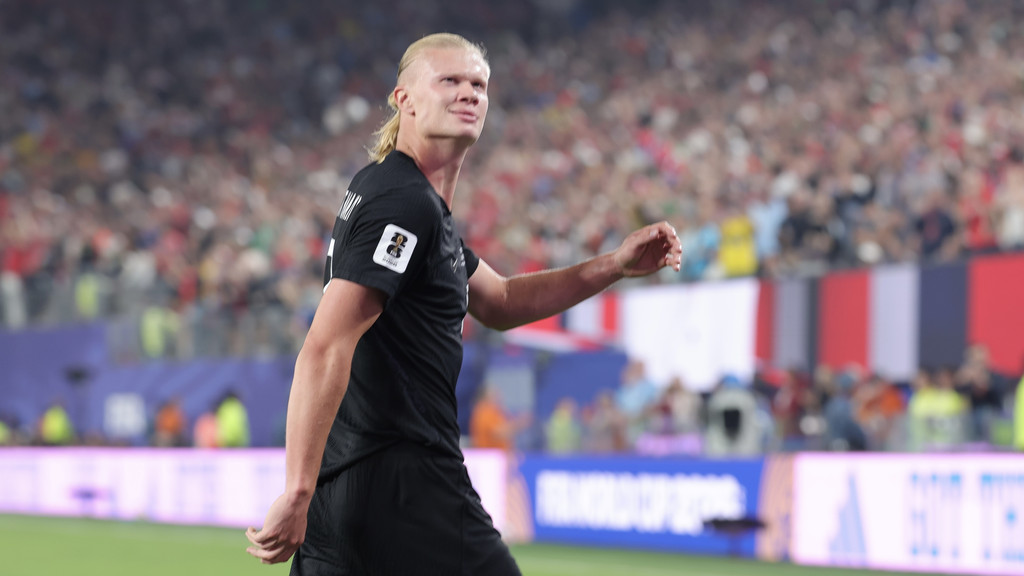 Erling Haaland, a male soccer player with blonde hair in a ponytail, wearing a black jersey, smiles while walking on a field with blurred crowds in the background.