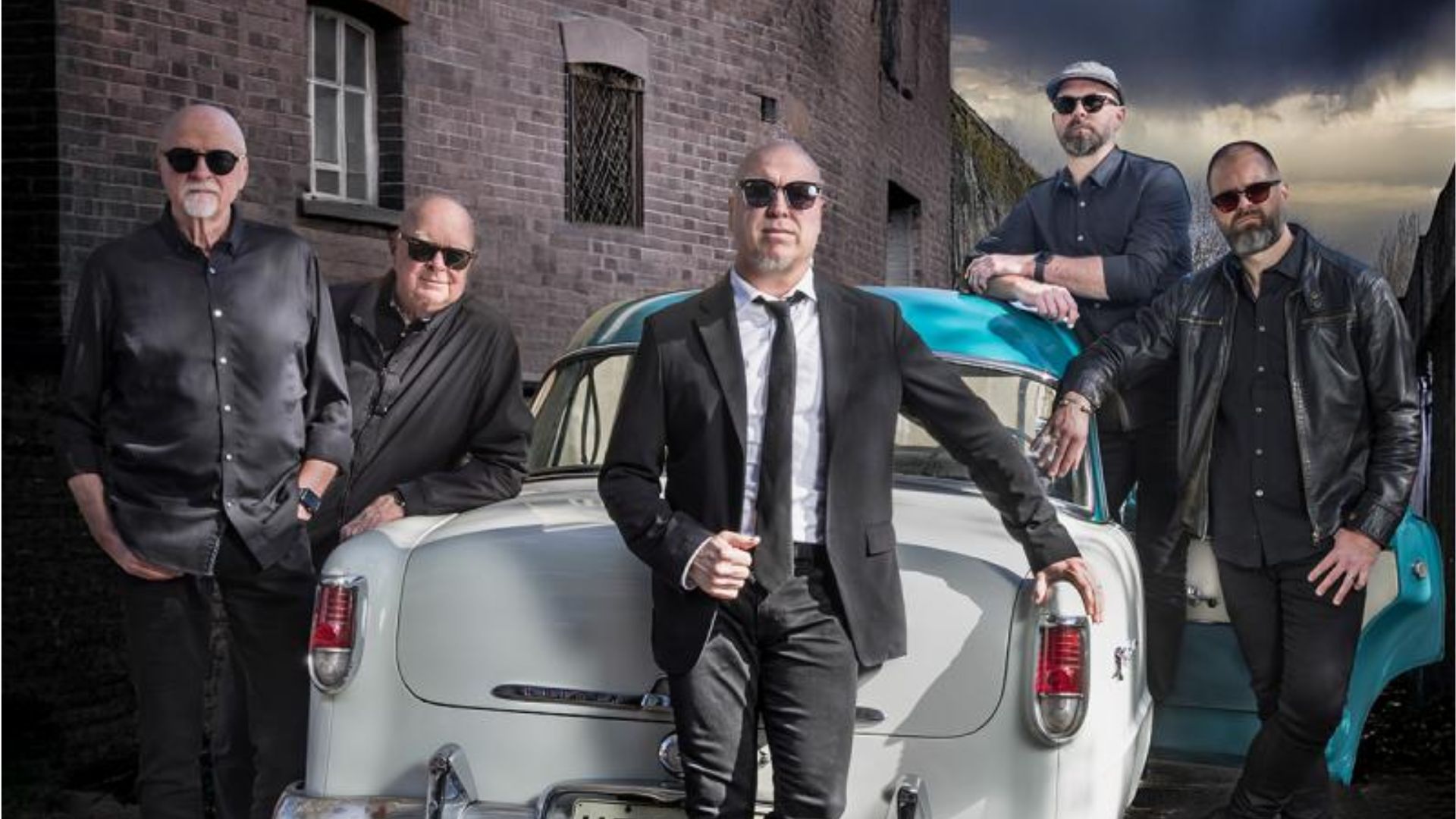 Five men in black attire and sunglasses pose around an old car with a light blue roof, in front of a brick building under a cloudy sky.