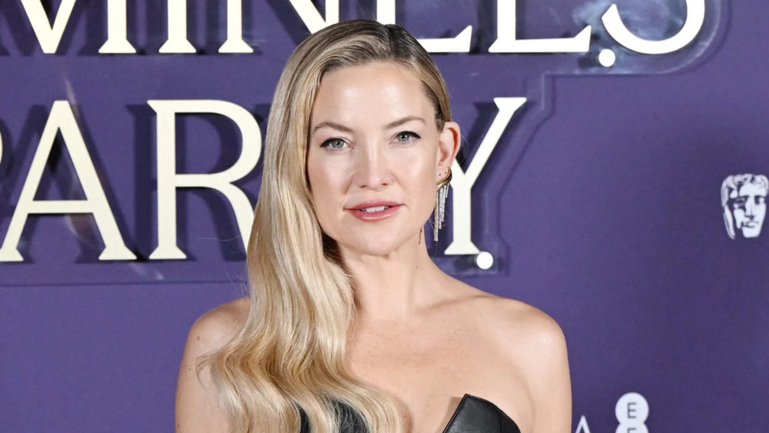 Kate Hudson, with long blonde wavy hair, looks directly at the camera, wearing a black strapless top and a sparkly drop earring, against a purple background with white text and a BAFTA logo.