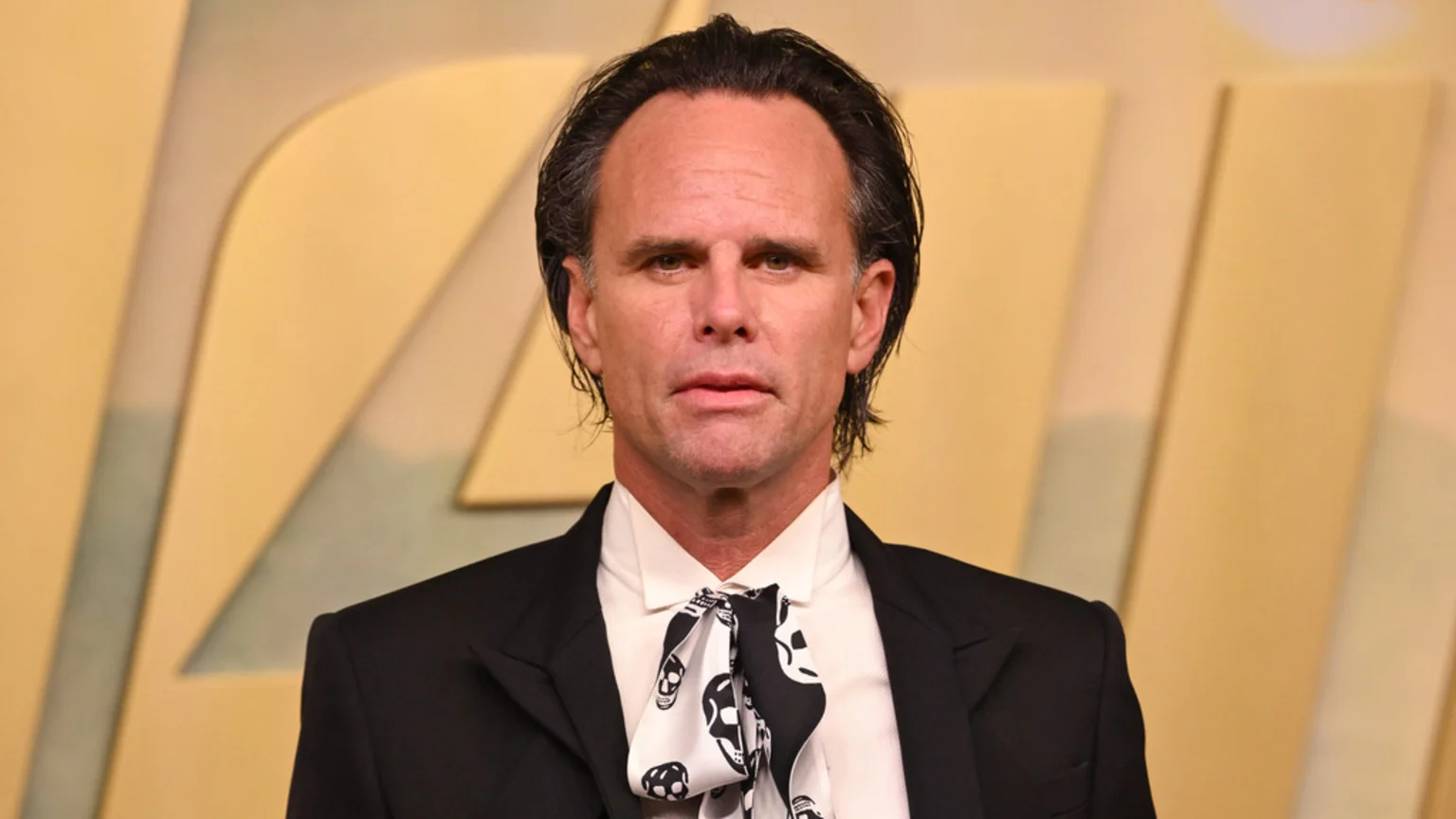 A man with dark, slicked-back hair, wearing a black suit jacket, a white shirt, and a black and white skull-patterned ascot, looks directly at the camera against a blurred light yellow background.