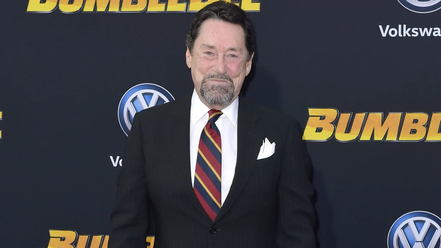 A smiling older man with a dark goatee, wearing a pinstriped suit, white shirt, and a red, navy, and gold striped tie, stands before a dark backdrop with "Bumblebee" and Volkswagen logos.