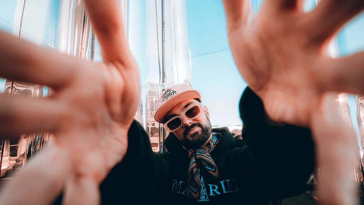 A bearded man in a pink cap and sunglasses looks at the wide-angle camera with both blurred hands reaching out, standing under a blue sky next to tall, reflective structures.