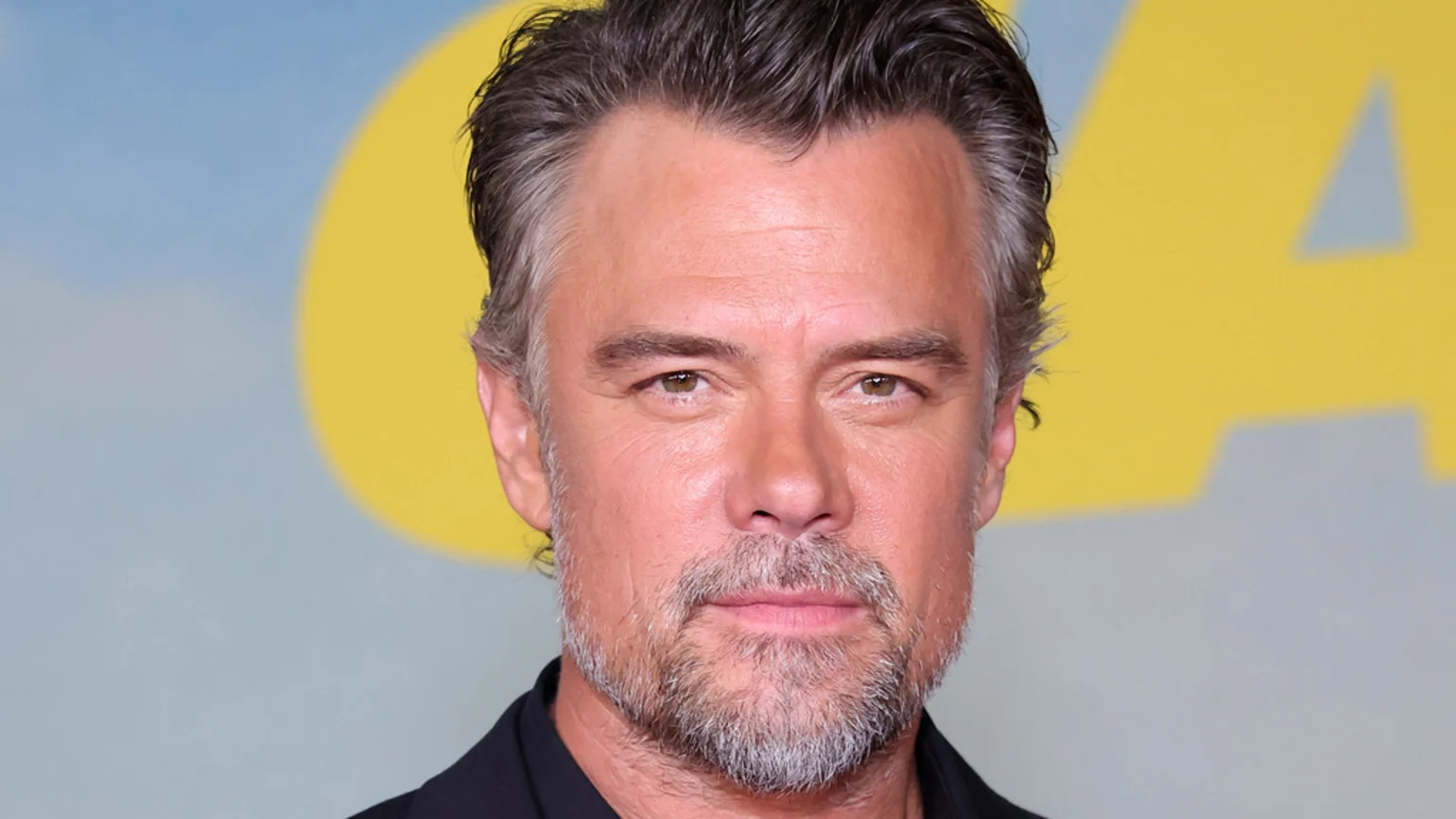 A close-up headshot of actor Josh Duhamel, with grey-streaked dark hair and a beard, looking directly at the camera against a light blue and yellow background.