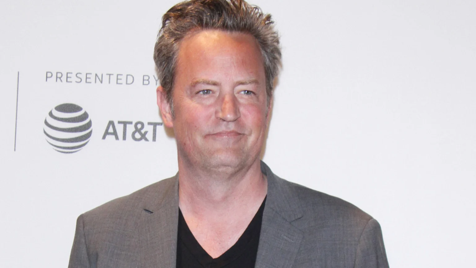 Matthew Perry, a middle-aged man with graying hair, smiles subtly while wearing a gray blazer over a black V-neck shirt, standing against a white wall with a "PRESENTED BY AT&T" logo.