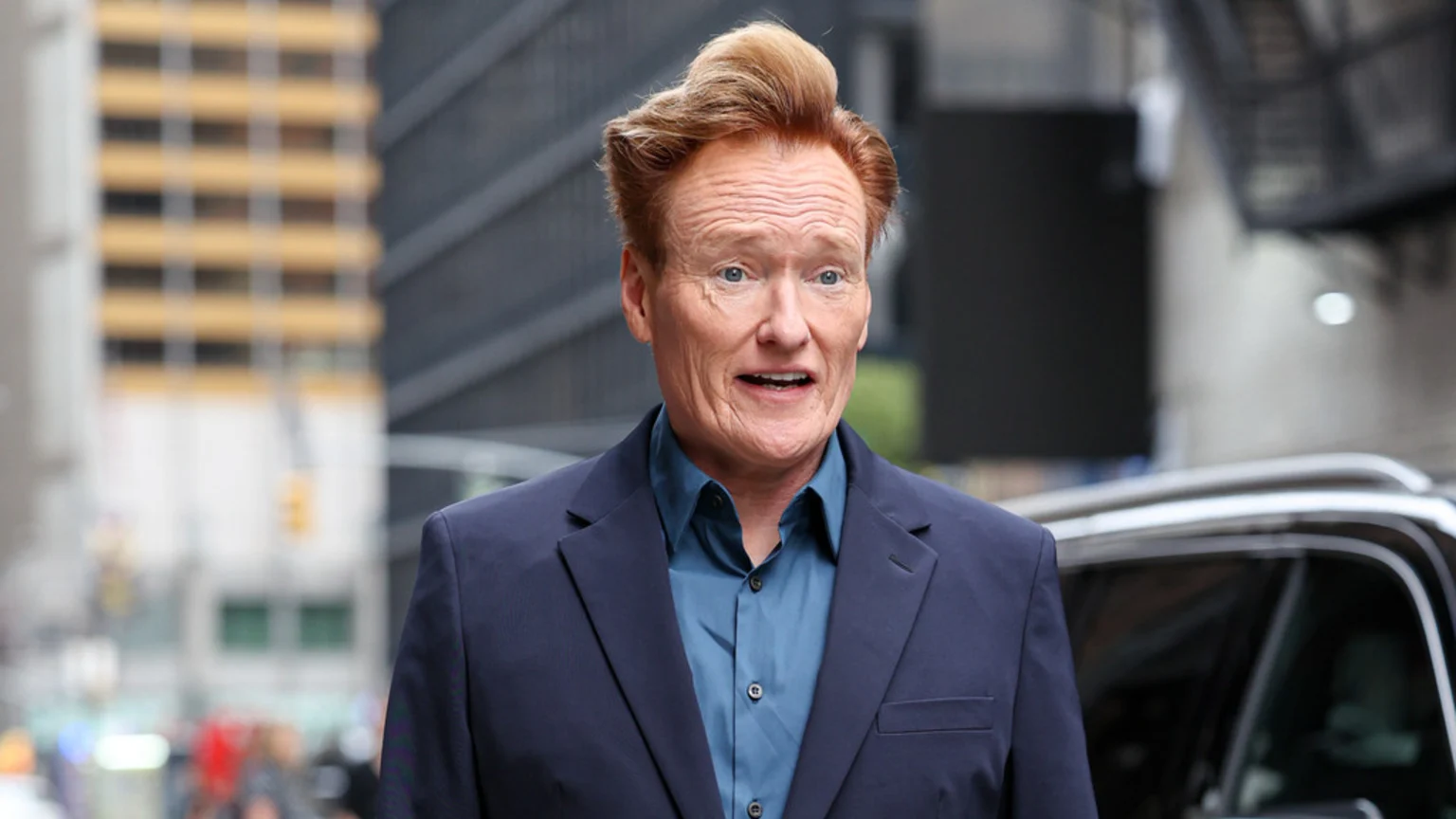 Conan O'Brien with his distinctive red hair, wearing a blue collared shirt and dark suit jacket, looks forward with a slightly open mouth in a blurred urban background.