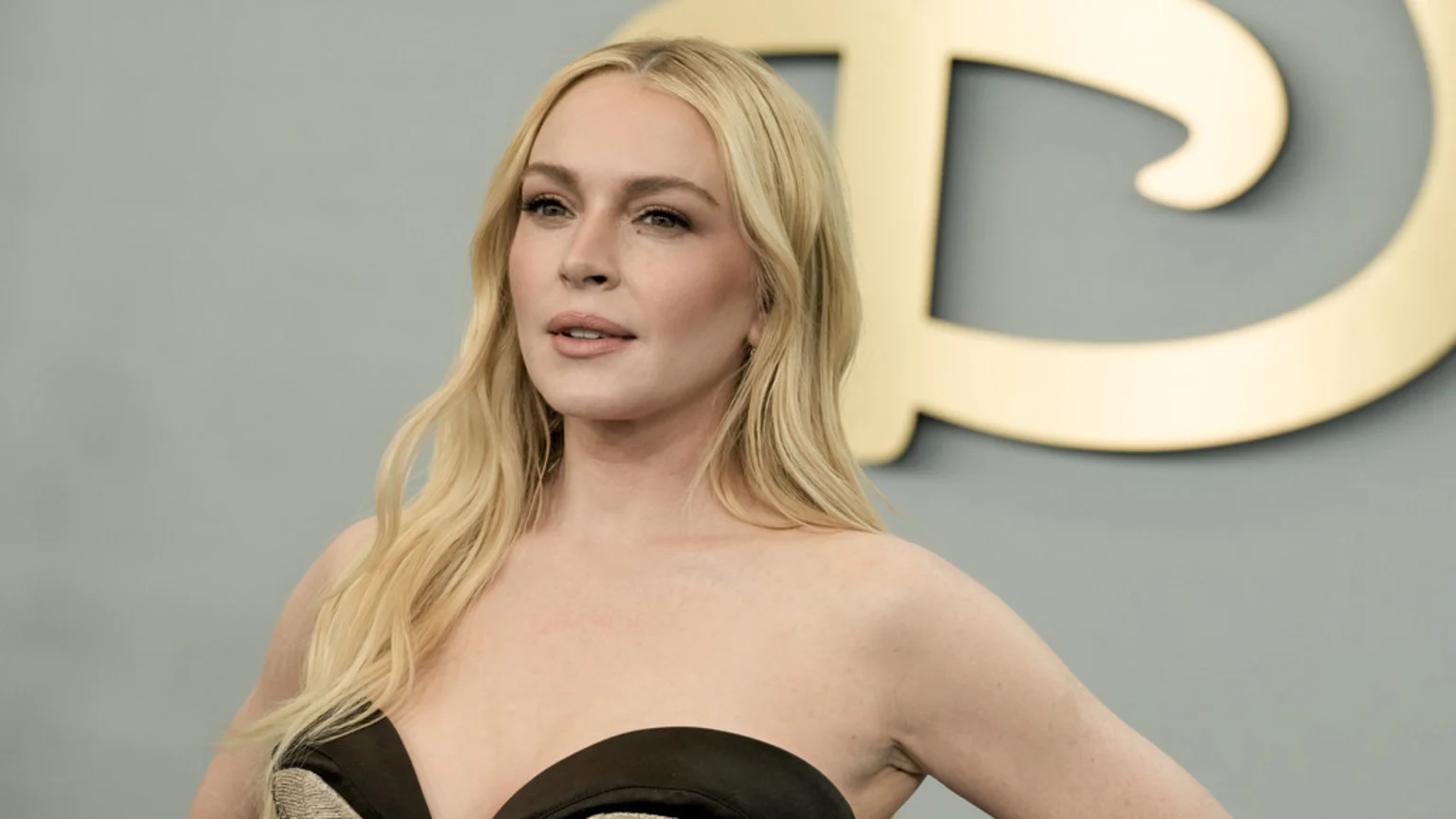 Lindsay Lohan with long wavy blonde hair and a strapless black top, looking to her left, against a blurred gray background with a gold 'D' logo.