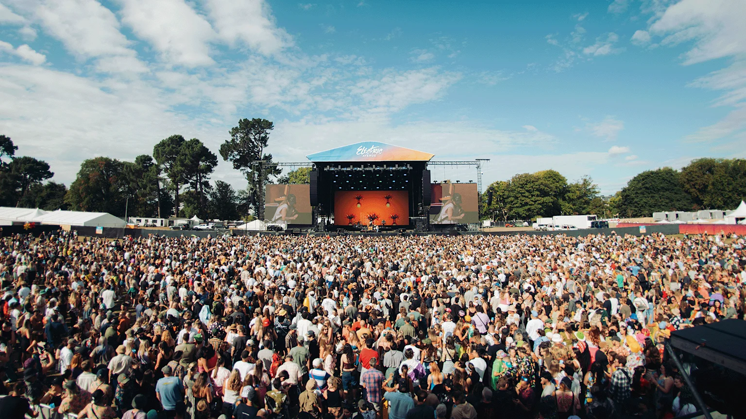 A wide view of a large crowd at an outdoor music festival, facing a stage with "Electric Avenue" branding, under a blue sky.
