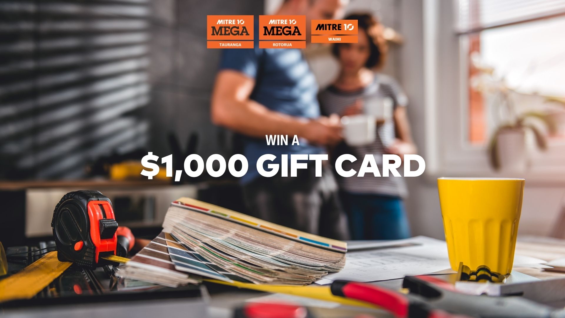 A tape measure, color swatches, tools, and blueprints are on a table, with blurry people in the background and overlay text 'WIN A $1,000 GIFT CARD' and Mitre 10 logos.