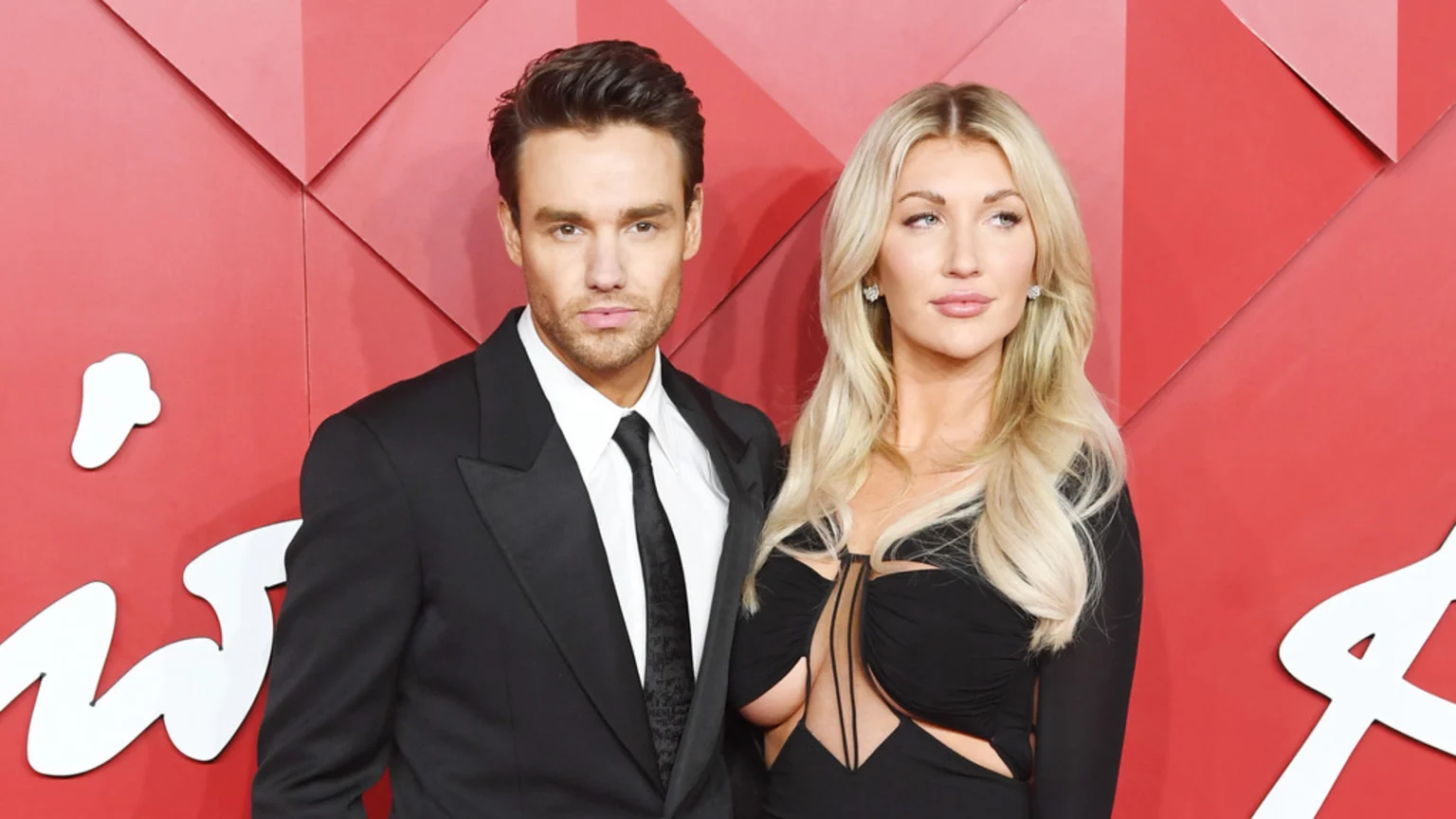 A man in a black suit and a woman in a black cutout dress with long blonde hair pose side-by-side against a geometric red background.