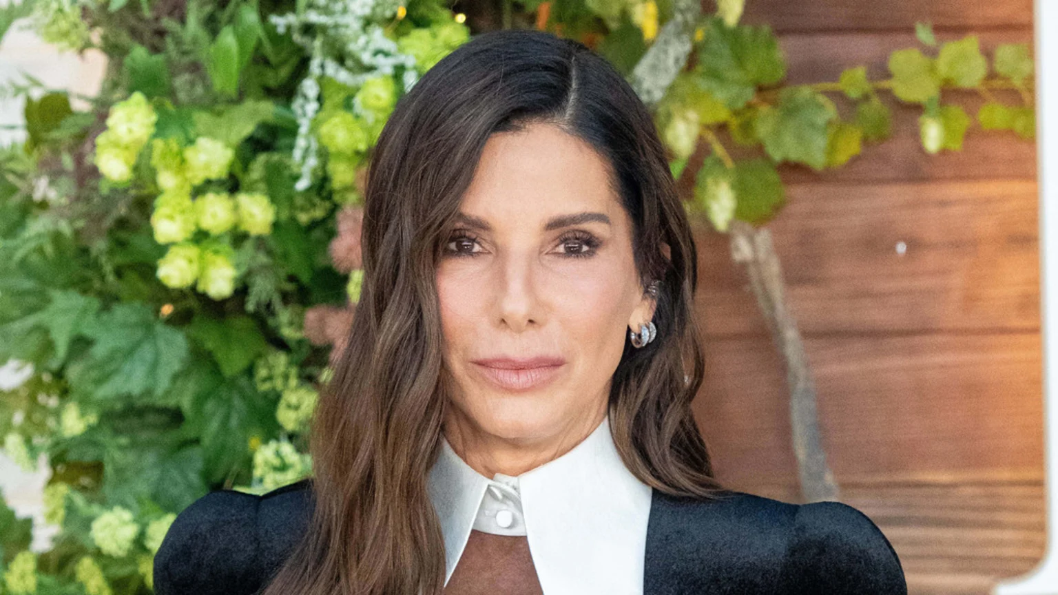 A woman with long brown hair and dark eyes wears a white collared shirt under a black jacket, against a blurred background of green foliage and a wooden wall.
