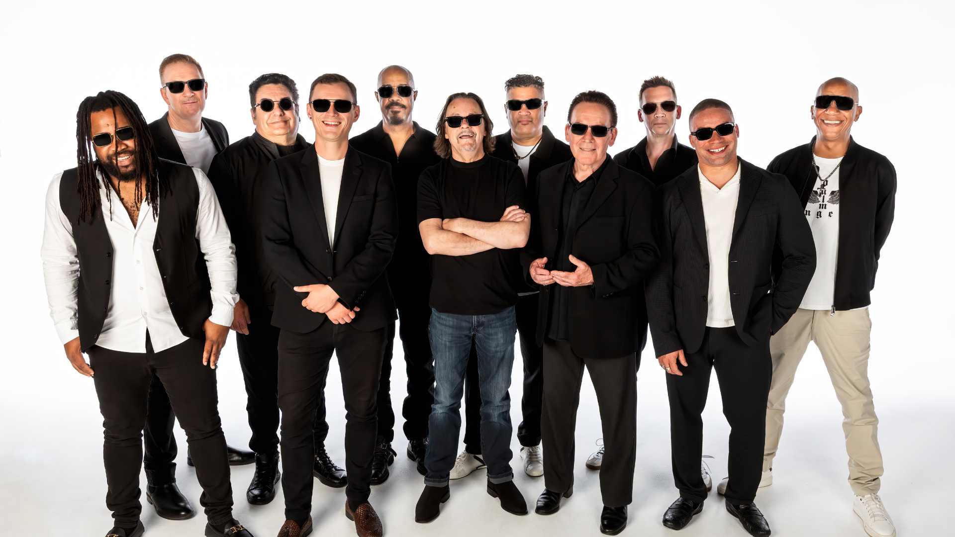 Eleven members of a band, mostly wearing black outfits and sunglasses, pose smiling for a group photo against a white background.