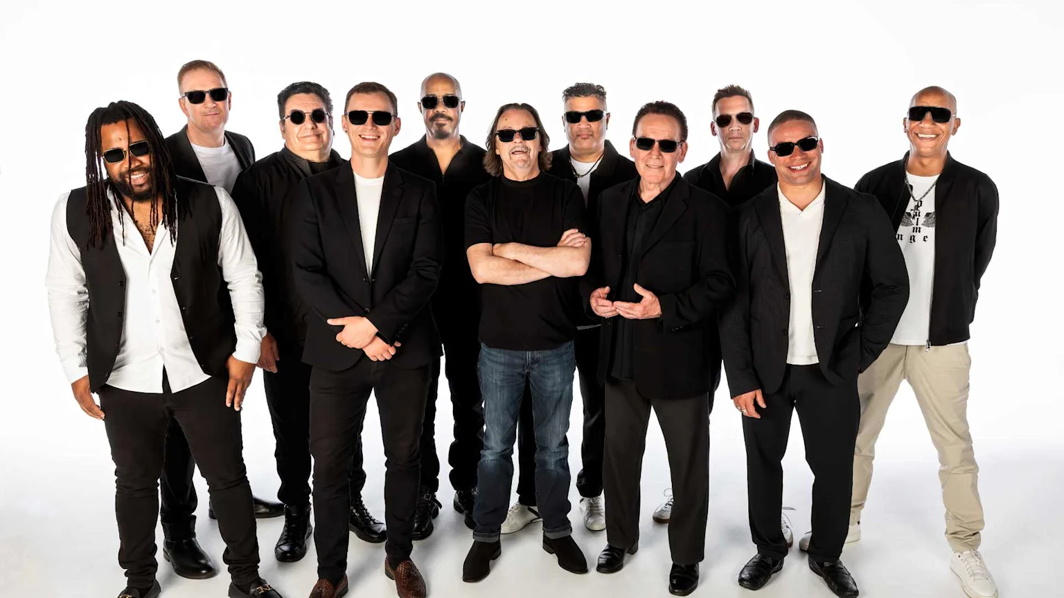 Eleven members of a band, mostly wearing black outfits and sunglasses, pose smiling for a group photo against a white background.