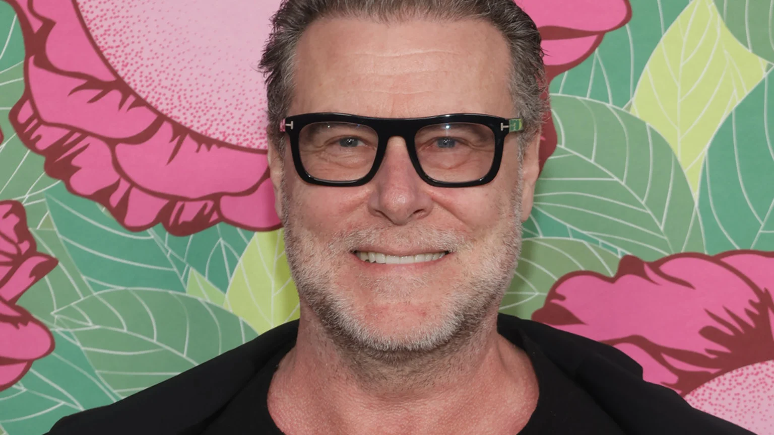Dean McDermott, a smiling man with graying hair and a beard, wears black glasses and a dark shirt against a vibrant background of pink flowers and green leaves.