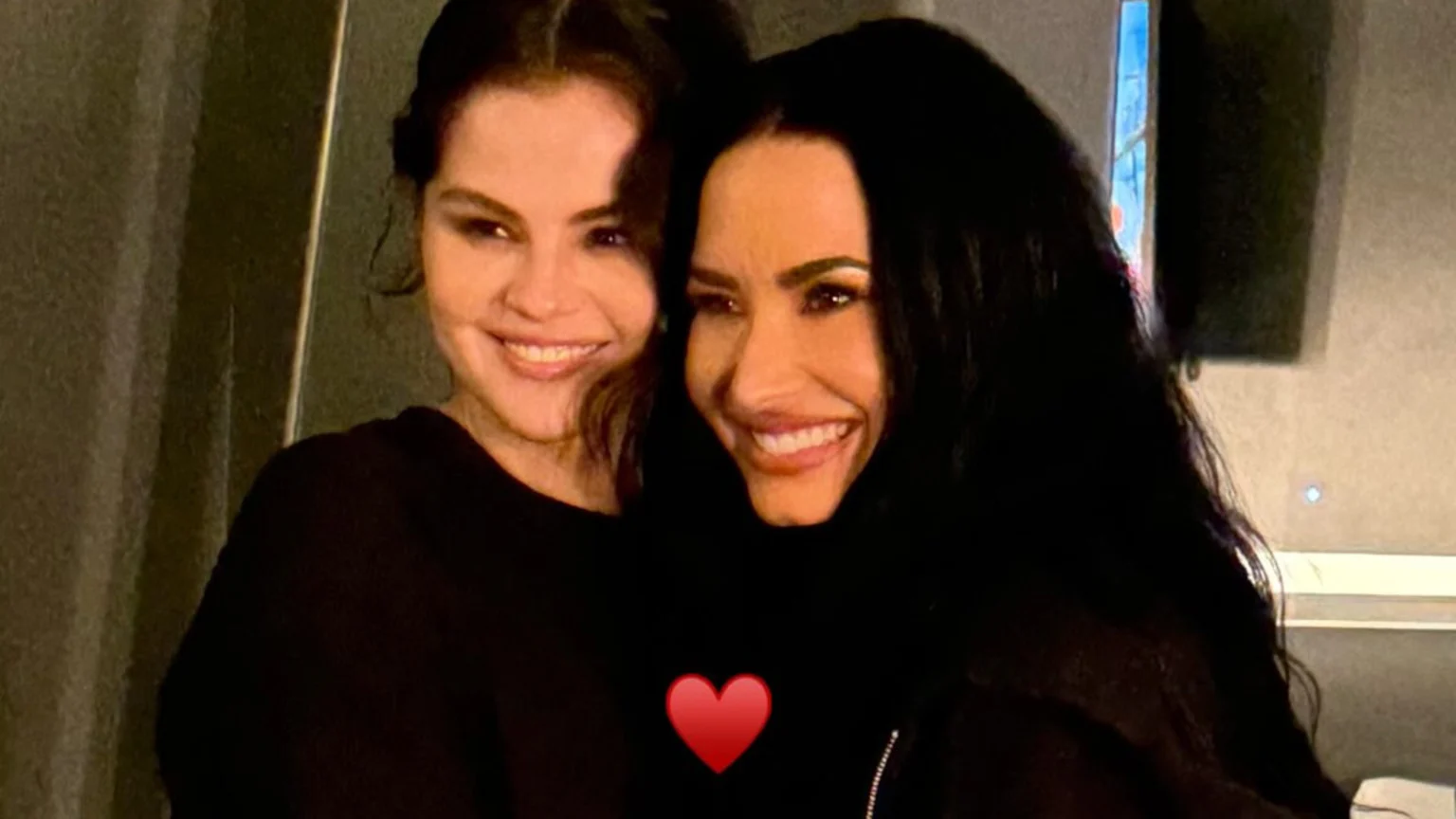 Selena Gomez and Demi Lovato smile broadly while posing closely together, with a red heart emoji covering their chests.