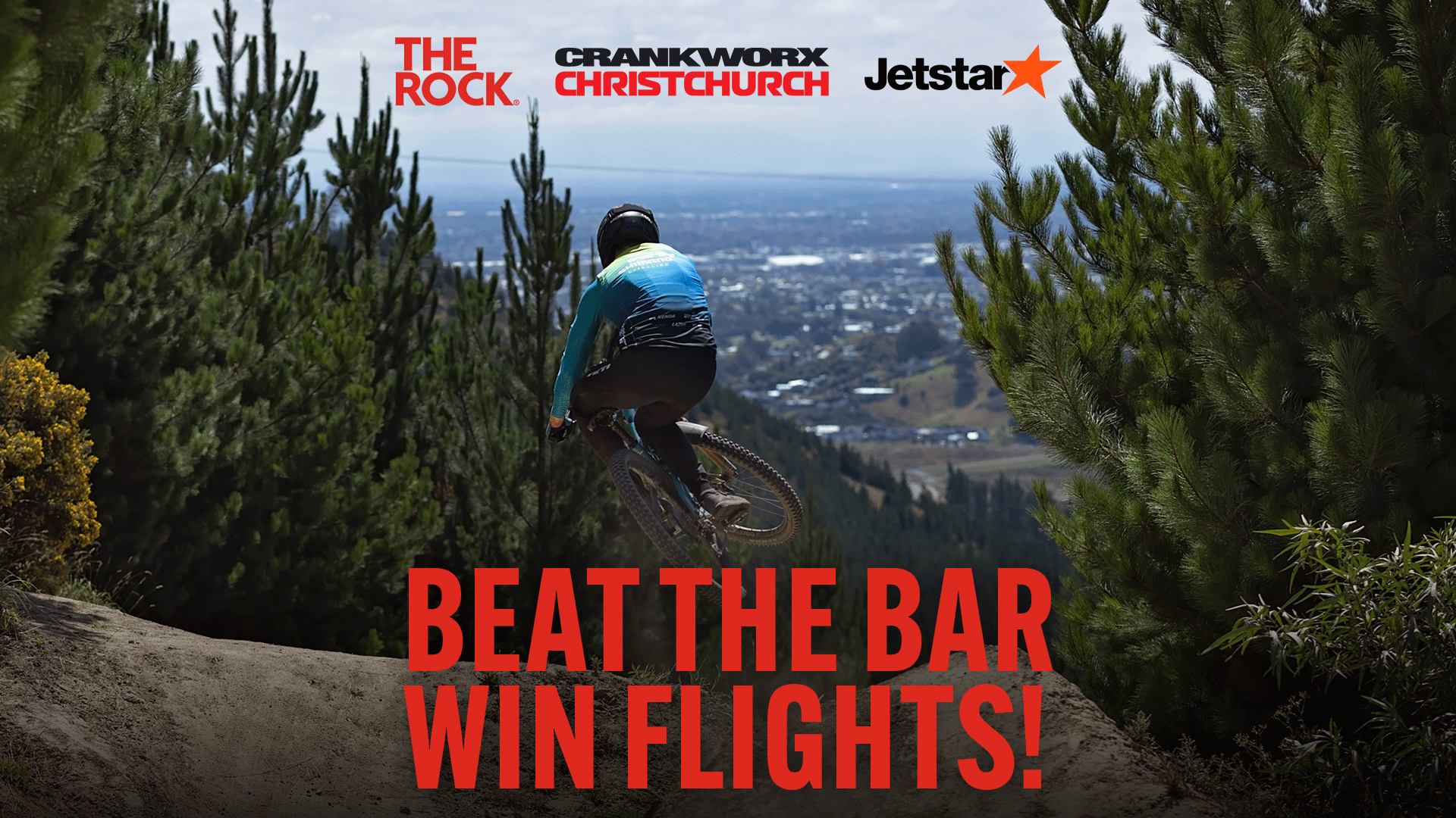 Action shot of a mountain biker wearing a blue and black jersey and full-face helmet jumping a dirt mound on a trail framed by pine trees. Cityscape visible in background. Features logos for The Rock, Crankworx Christchurch, and Jetstar, plus 'Beat The Bar Win Flights!' text.