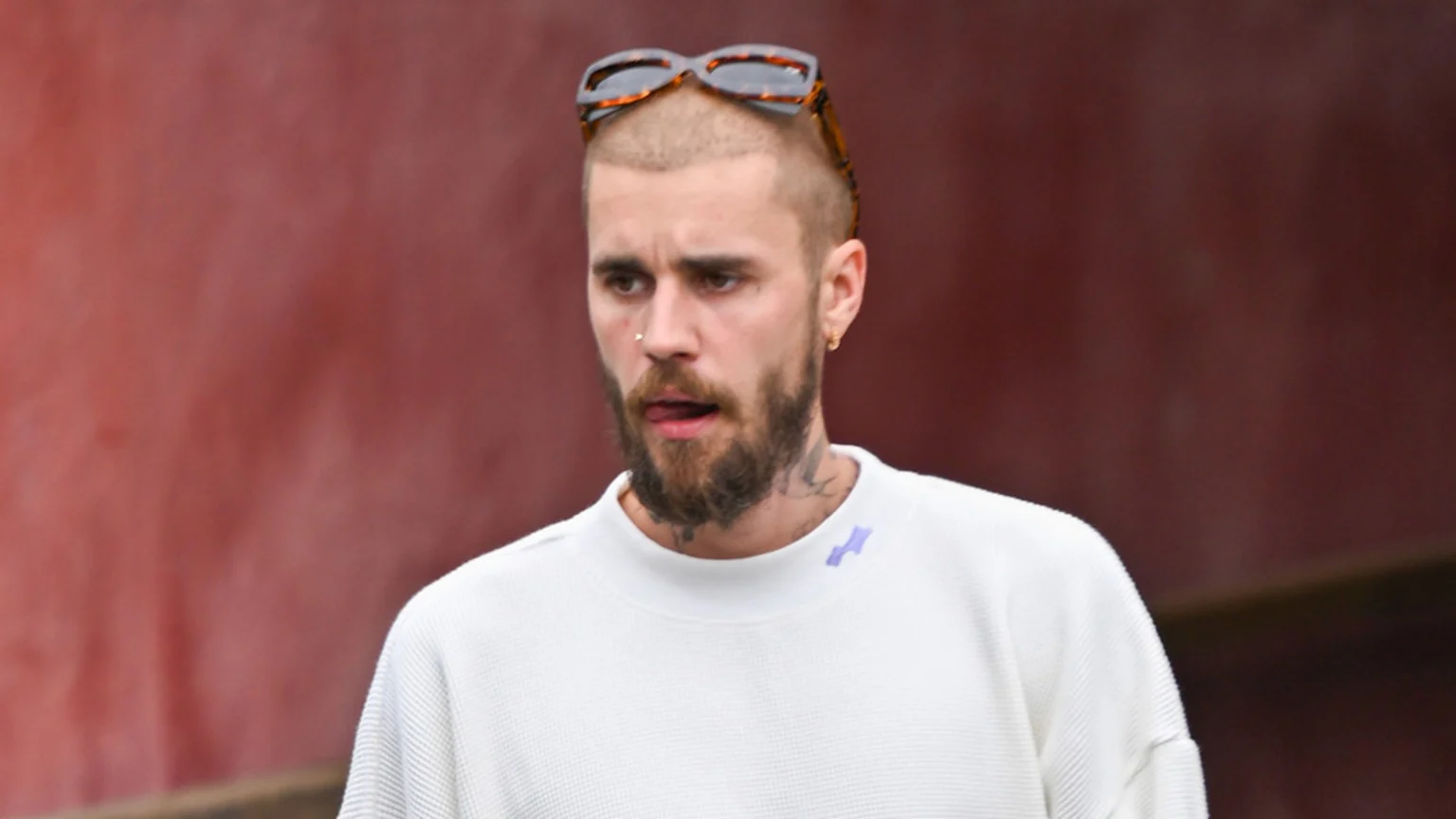 Justin Bieber with a shaved head, beard, sunglasses on his head, nose ring, and neck tattoos, wears a white waffle-knit shirt and sticks his tongue out slightly against a reddish-brown background.
