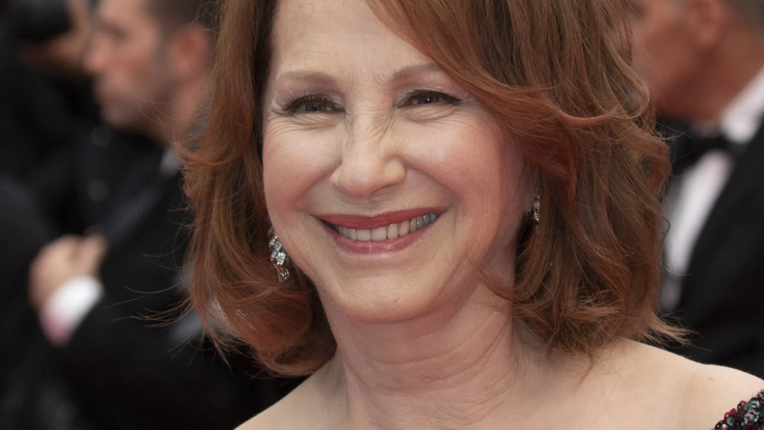 A smiling woman with reddish-brown, wavy, shoulder-length hair, visible wrinkles around her eyes, and sparkly earrings, against a blurred background of people in formal attire.