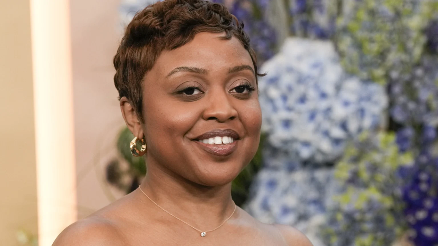 Close-up of Quinta Brunson smiling, with short brown hair, gold hoop earrings, and a delicate necklace, against a blurred background of blue and green flowers.