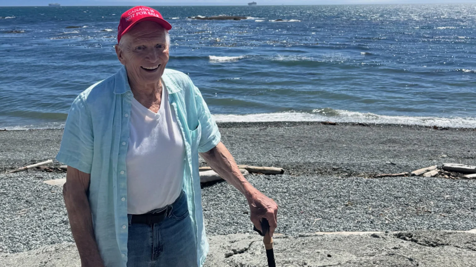An elderly man in a red "CANADA IS NOT FOR SALE" hat, light blue shirt, and jeans smiles on a sunny pebbly beach by the ocean, holding a walking stick.