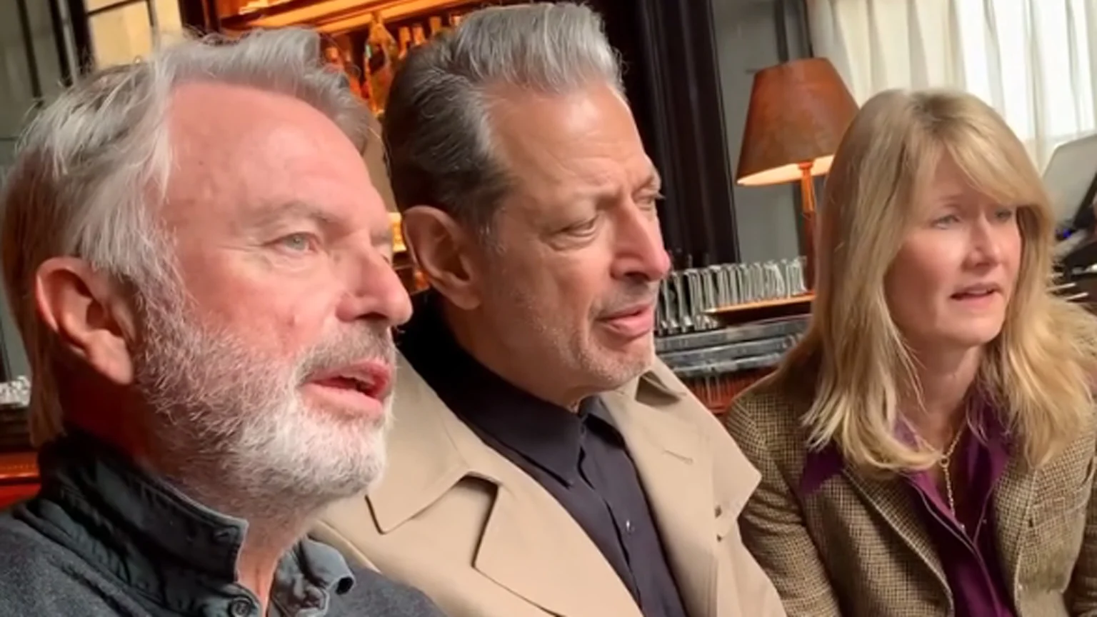 Sam Neill with a grey beard, Jeff Goldblum in a tan trench coat, and Laura Dern with blonde hair are seated indoors, looking right.