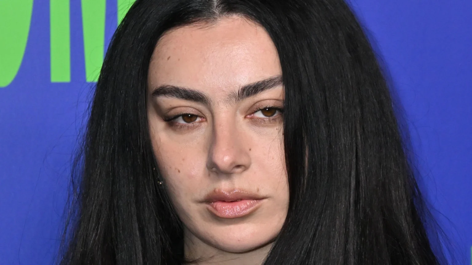 A close-up of Charli XCX with dark hair parted down the middle, thick eyebrows, and brown eyes, gazing slightly down against a vibrant blue and green background.