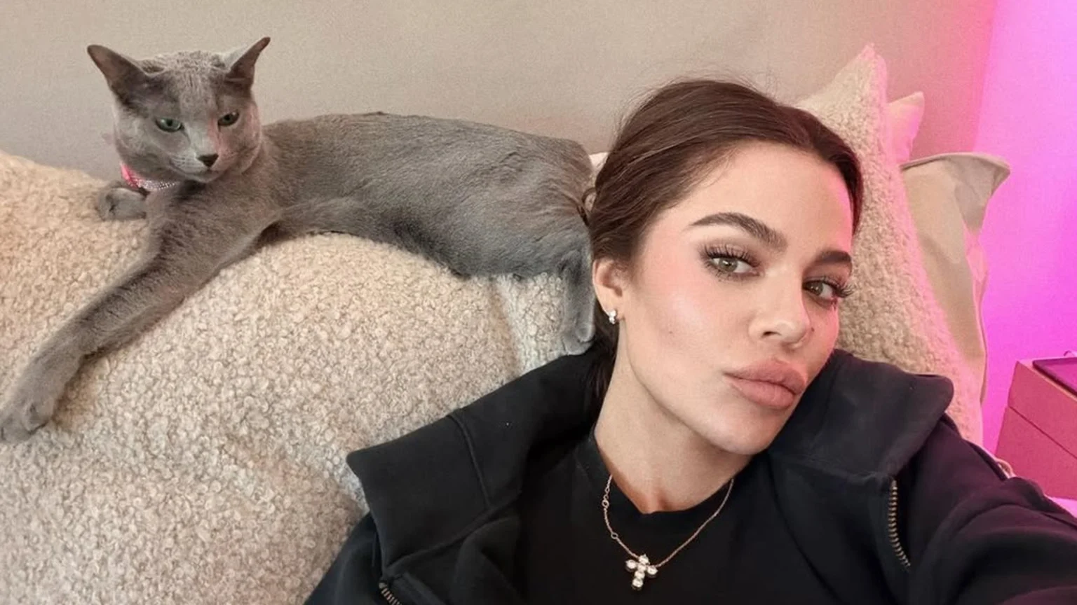 Khloé Kardashian takes a selfie next to her grey cat wearing a pink sparkly collar, both resting on a light-colored, textured surface with a soft pink light in the background.
