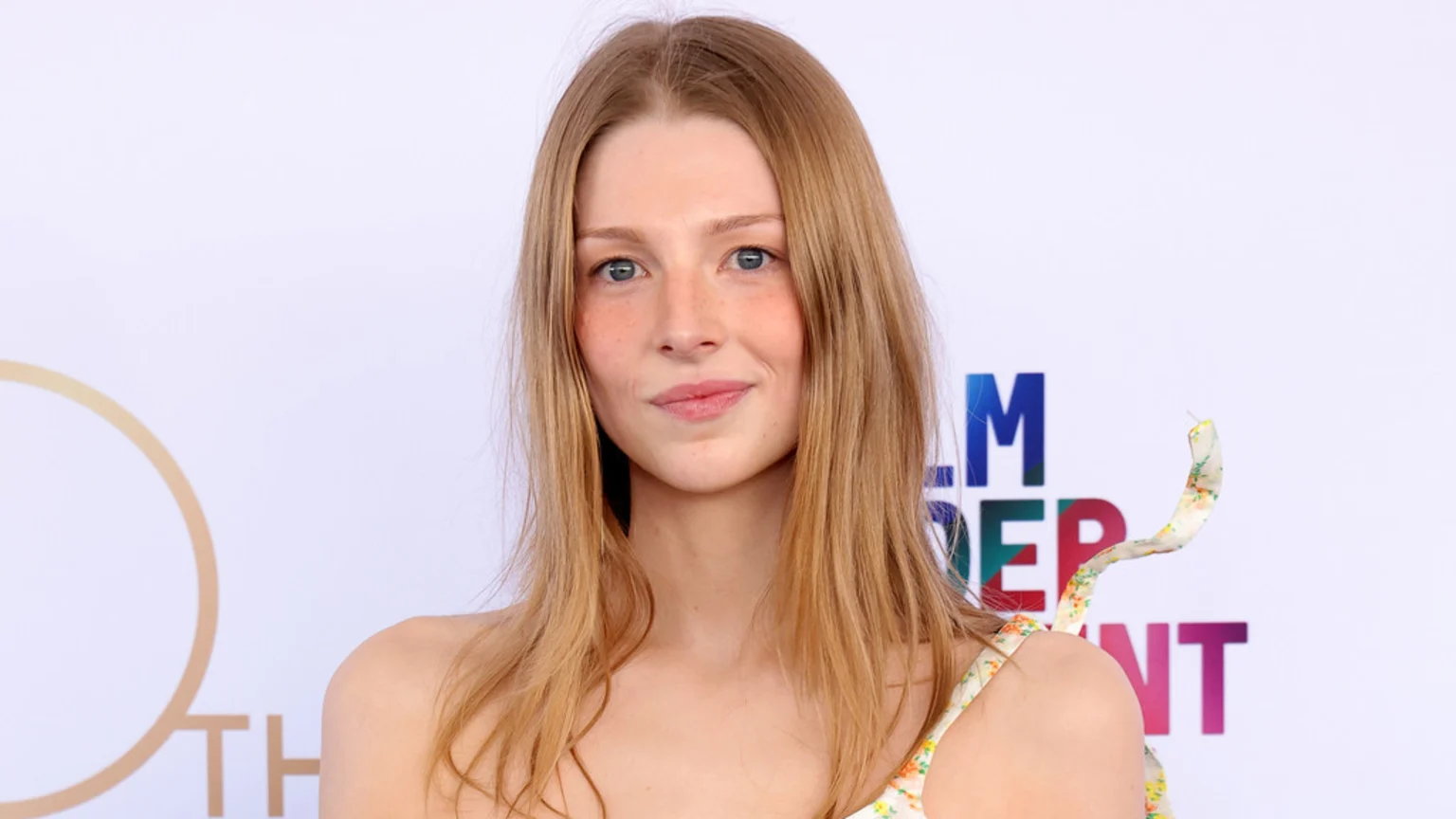 A young woman with light brown hair, blue eyes, and freckles smiles slightly, wearing a floral strap top against a white background with blurred logos.