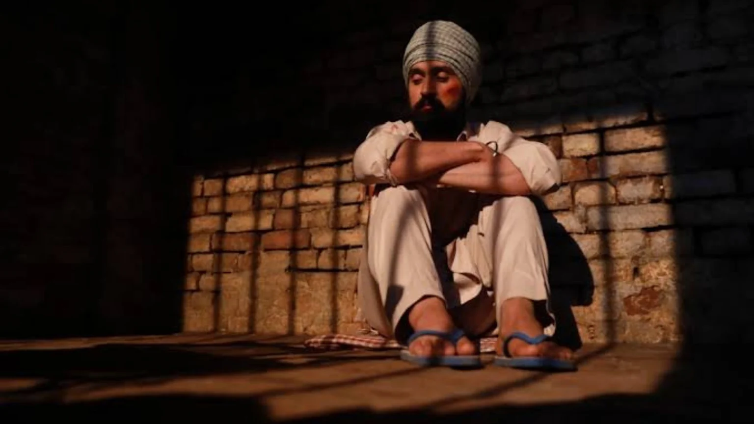 A man in a turban and beard, with a red mark on his cheek, sits with crossed arms on the floor of a dark, brick-walled room, where light casts shadows of prison bars.