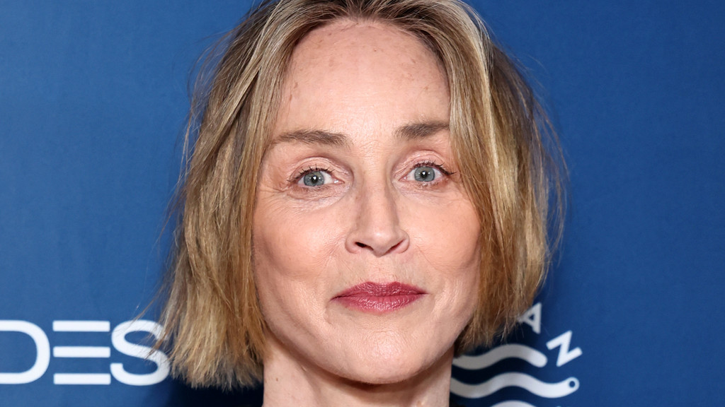 Actress Sharon Stone looks directly at the camera in a close-up portrait, with blonde bob-length hair, light blue eyes, and reddish lipstick, against a blue background.
