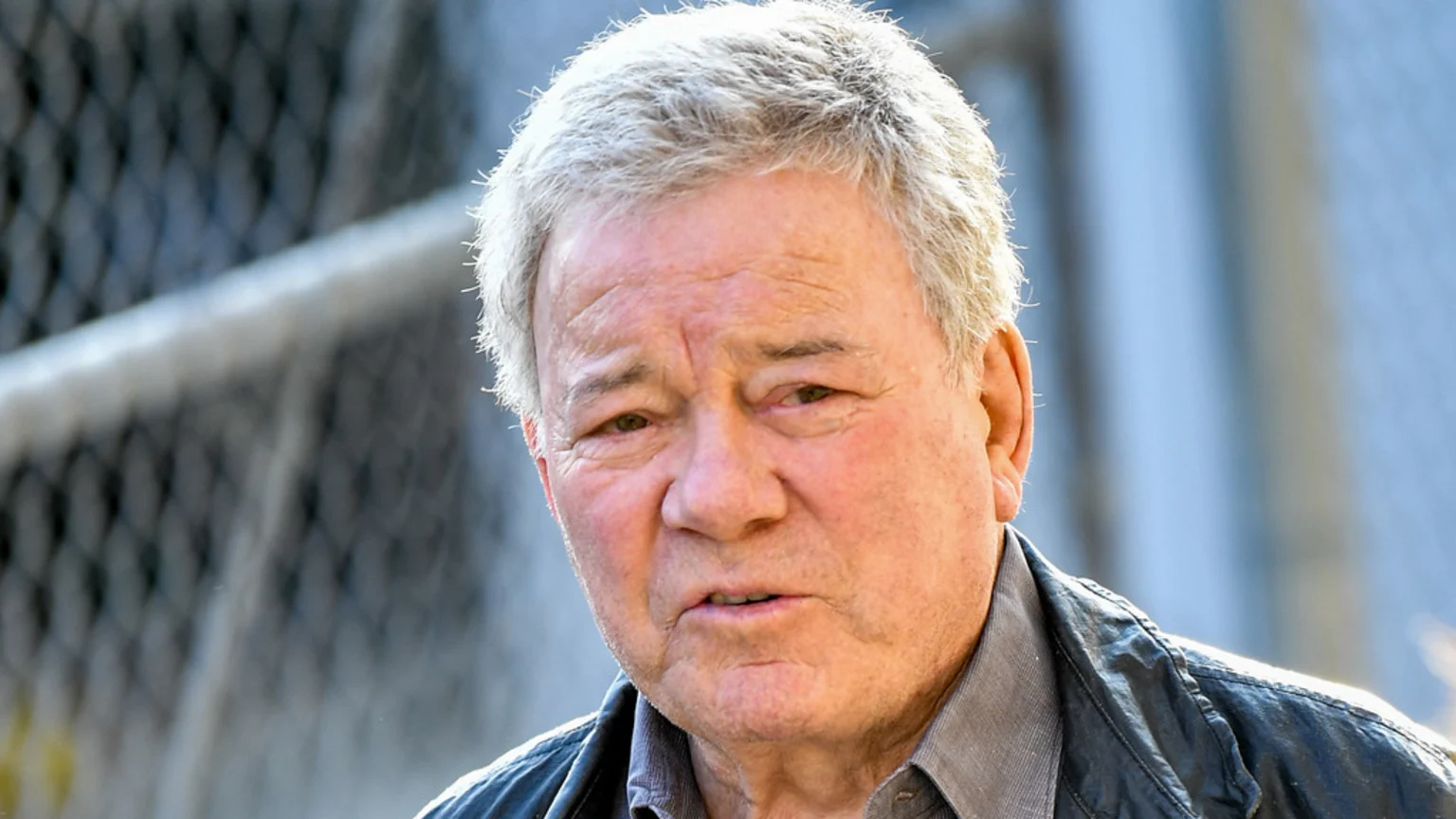A close-up of actor William Shatner, an older man with gray hair, looking forward with his mouth slightly open, wearing a dark jacket over a gray collared shirt.