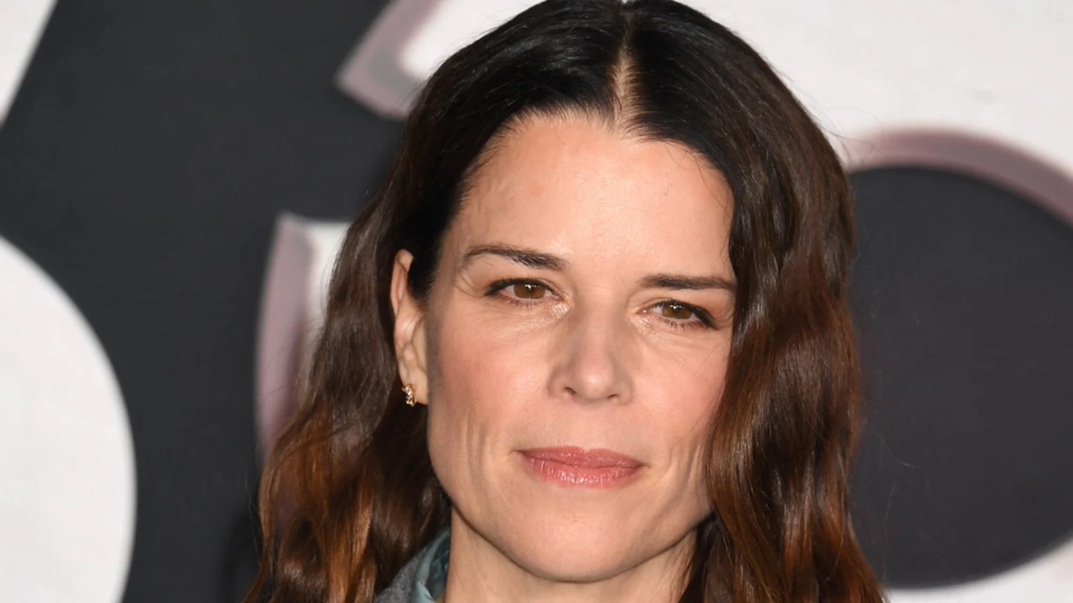 A close-up headshot of Neve Campbell, with long dark brown wavy hair, light brown eyes, and a slight smile, wearing a light-colored top, against a blurred background.