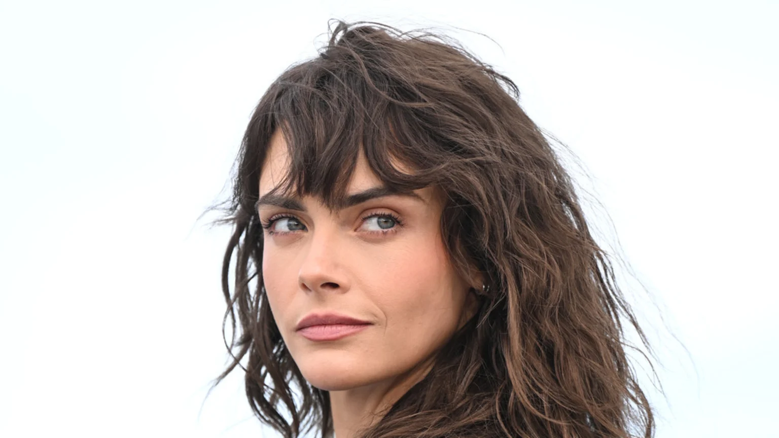 A close-up portrait of a woman with dark, wavy hair and bangs, light eyes, and subtle makeup, looking slightly to her right against a plain white background.