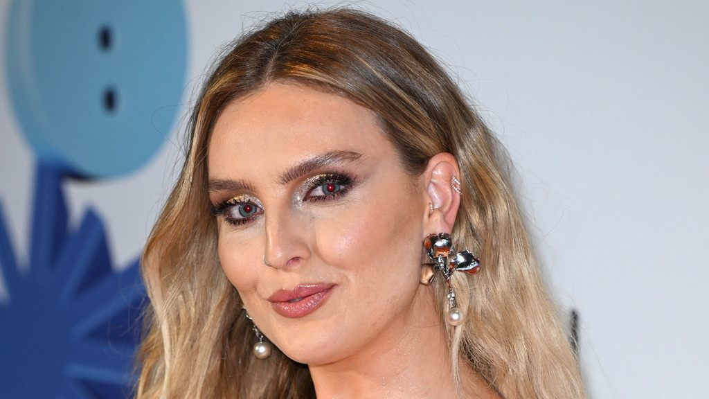 Perrie Edwards smiles slightly, showcasing long wavy blonde hair, striking glittery eye makeup with red-tinted pupils, a nose ring, and intricate silver and pearl dangle earrings.