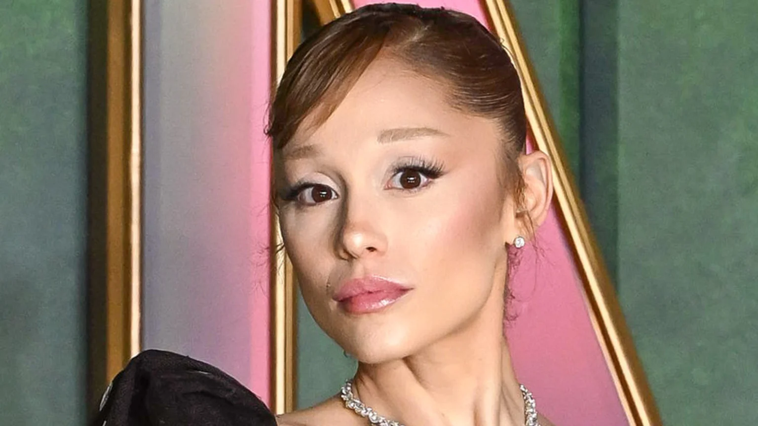 Close-up of Ariana Grande looking directly at the camera, with light brown hair, prominent eye makeup, pink glossy lips, and wearing a diamond earring and necklace against a blurred pink, gold, and green background.