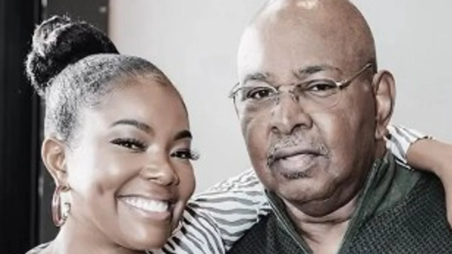 Smiling woman with hair in a bun embraces an older bald man in glasses and a dark sweater.