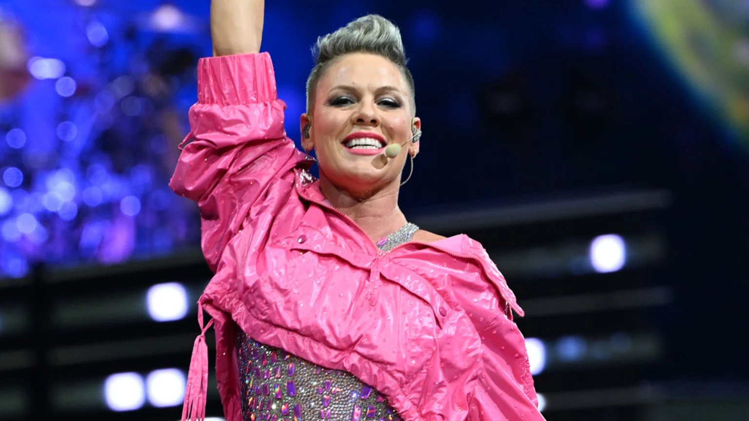Singer Pink smiles at the camera, wearing a shiny pink jacket and sparkly top, with her arm raised during a performance.