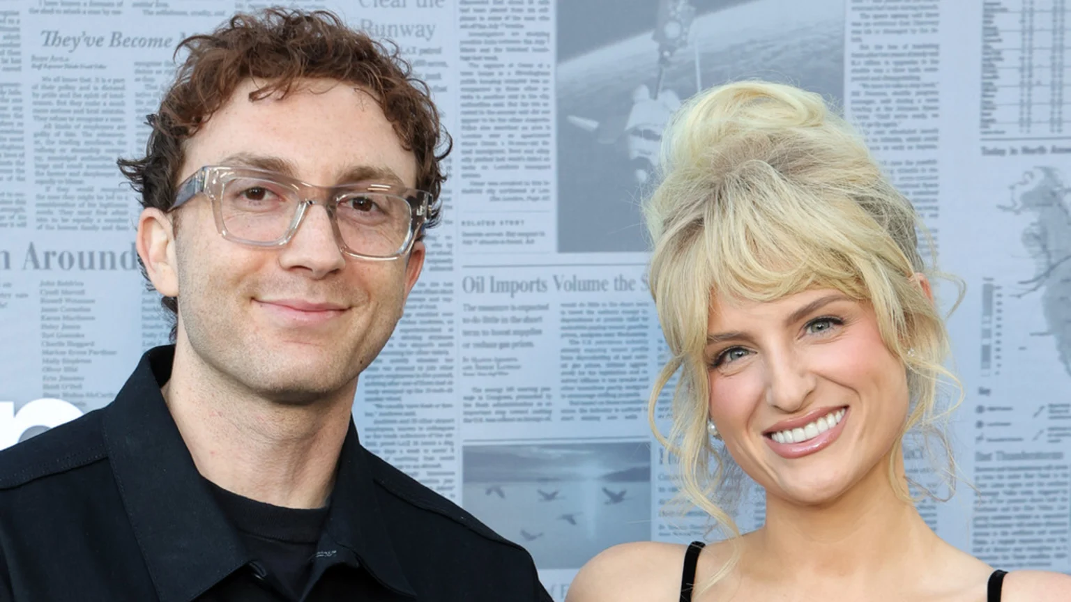 A man with curly brown hair and clear glasses smiles next to a broadly smiling woman with blonde hair and bangs against a document background.