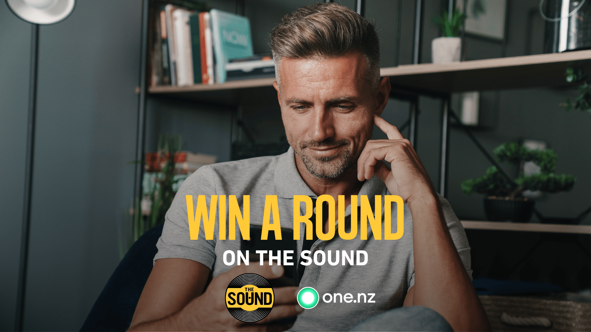 A man in a grey polo shirt looks at his smartphone with the text "WIN A ROUND ON THE SOUND" and logos for "THE SOUND" and "one.nz" superimposed on the image.