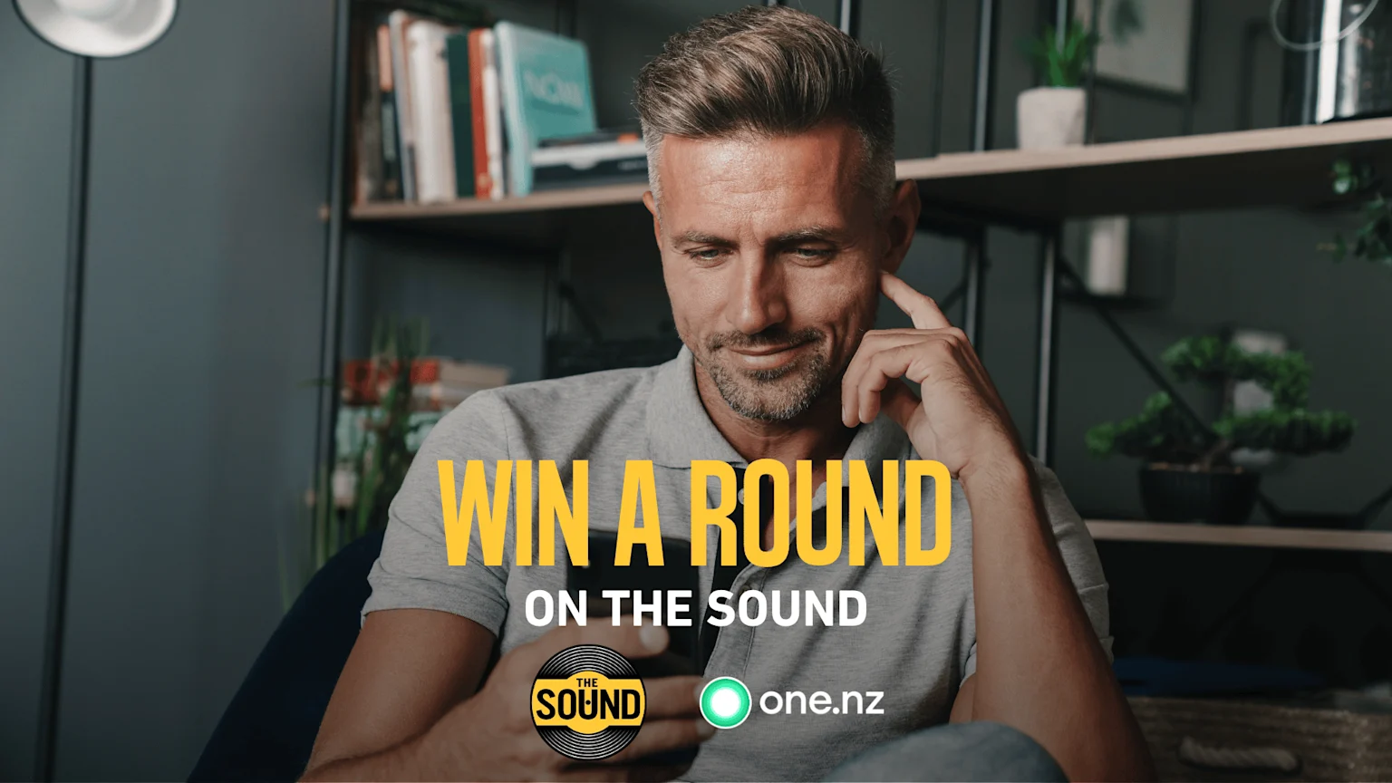 A man in a grey polo shirt looks at his smartphone with the text "WIN A ROUND ON THE SOUND" and logos for "THE SOUND" and "one.nz" superimposed on the image.