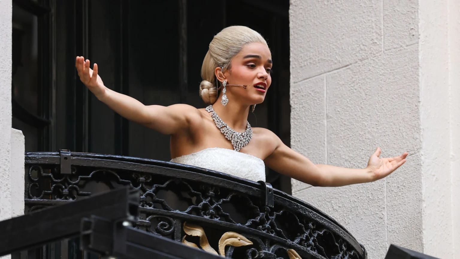A woman with blonde hair, wearing a white strapless gown and sparkling jewelry, sings passionately on a balcony with outstretched arms.