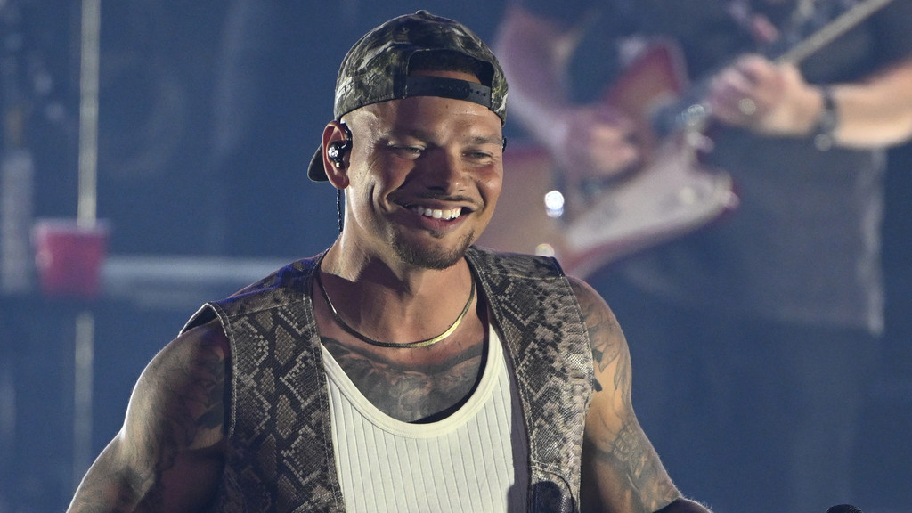 Kane Brown smiles brightly, wearing a camouflage cap backwards, in-ear monitors, a snake-print vest over a white tank top, a gold chain, and visible arm and chest tattoos.