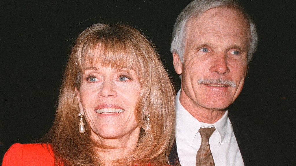 Jane Fonda, with blonde hair and a red top, smiles broadly next to Ted Turner, who has a gray mustache, gray hair, and wears a white shirt and patterned tie.