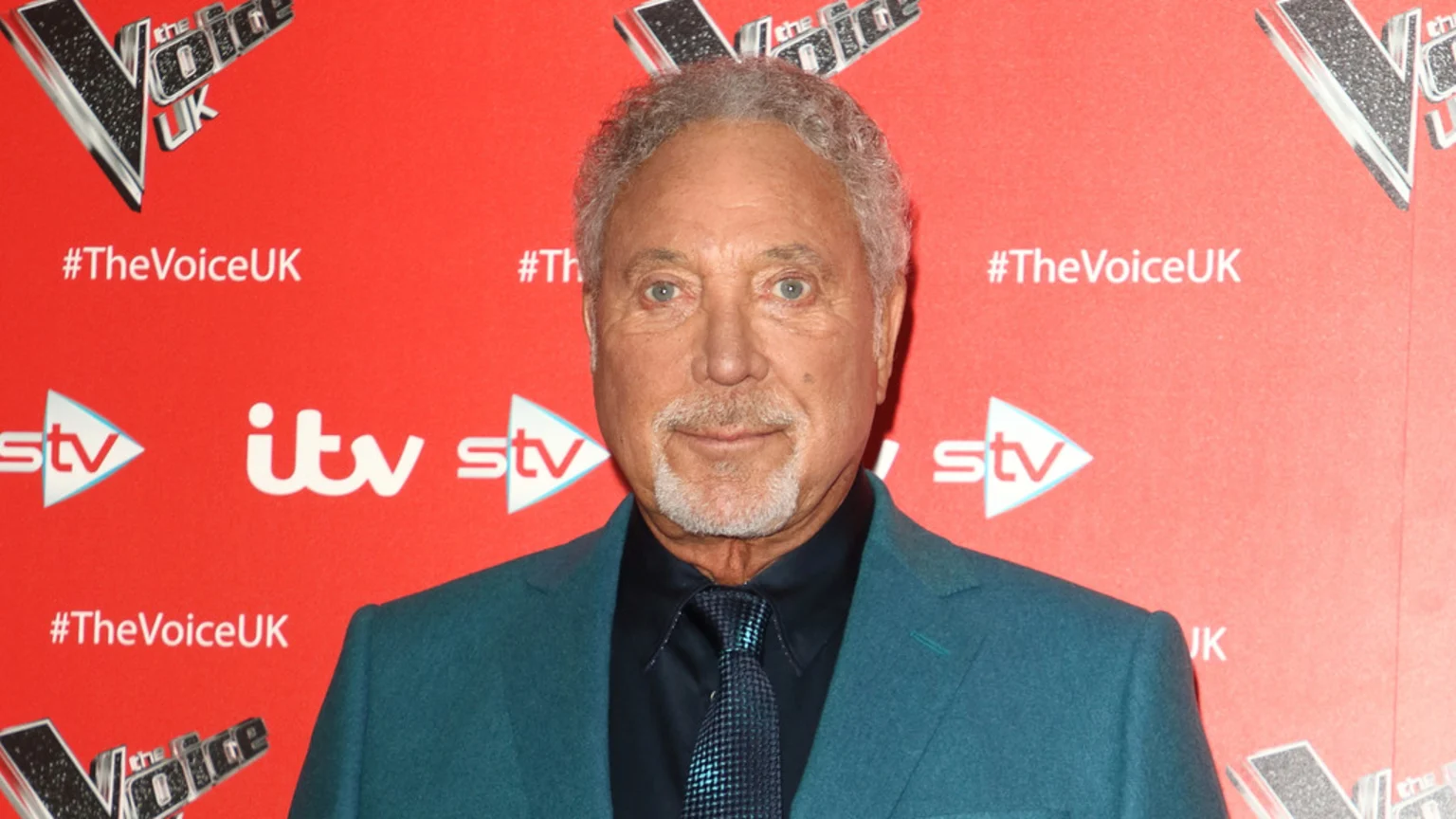 Tom Jones, an older man with grey hair and goatee, looks directly at the camera in a teal suit jacket against a red backdrop featuring "The Voice UK," "itv," and "stv" logos.