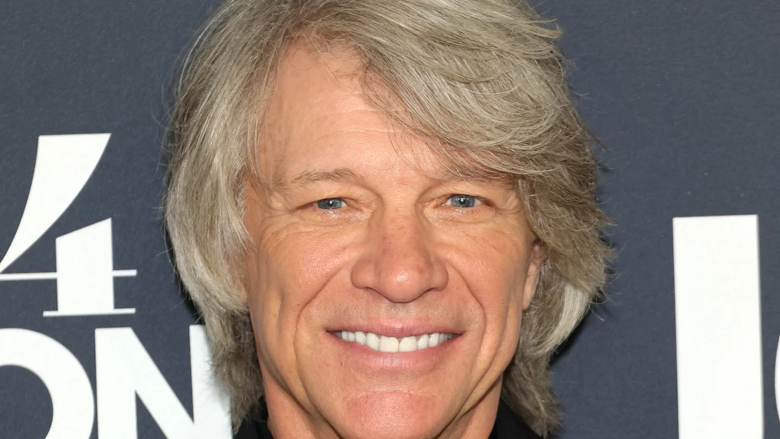 A close-up of singer Jon Bon Jovi, a man with long, shaggy grey hair and blue eyes, smiling against a dark background with partially visible white text.