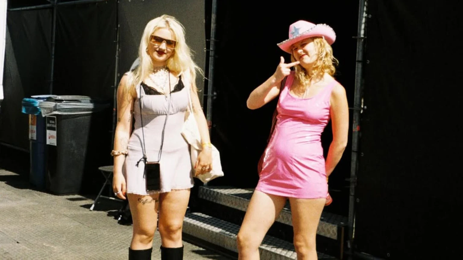 Two blonde women standing outdoors; one on the left wears a light dress and sunglasses, the other on the right wears a pink dress and a pink cowboy hat, making a hand gesture.