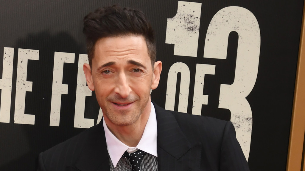 Adrien Brody smiles subtly, wearing a dark suit, white shirt, and polka-dot tie, with distressed white lettering on a black background behind him.