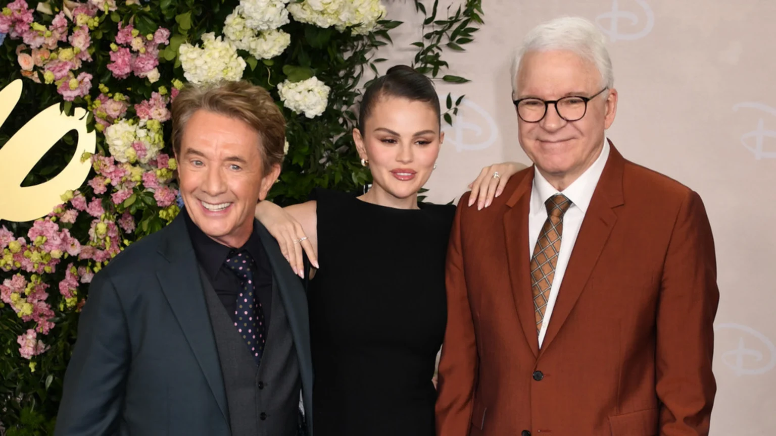Martin Short, Selena Gomez, and Steve Martin smile together at an event, with Martin Short in a dark suit, Gomez in a black dress, and Martin in a brown suit.