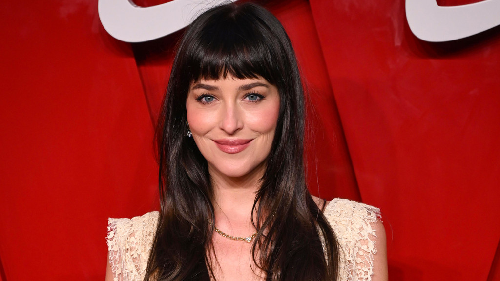 Dakota Johnson with dark hair and bangs and blue eyes smiles softly, wearing a lace top and delicate necklace, against a vibrant red background.